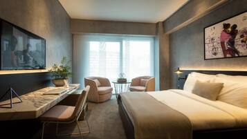 Une chambre d’hôtel moderne avec un grand lit, un bureau avec une chaise, une petite table avec deux chaises, une plante en pot et des œuvres d’art accrochées aux murs.