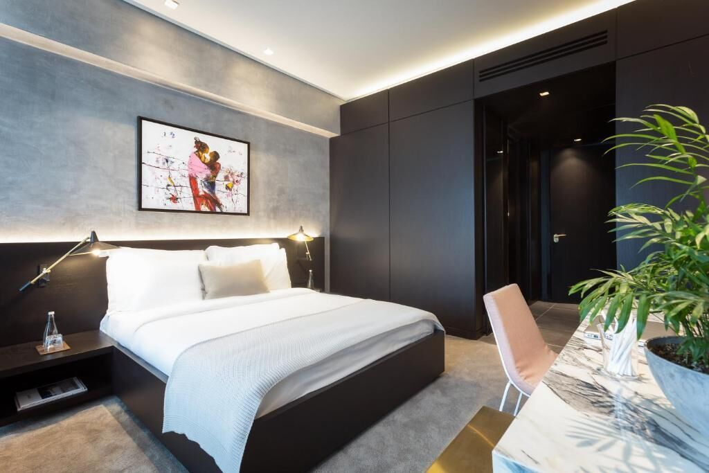 A modern hotel room with a large bed, bedside tables, a desk with a chair, a potted plant, and a framed artwork on the wall.