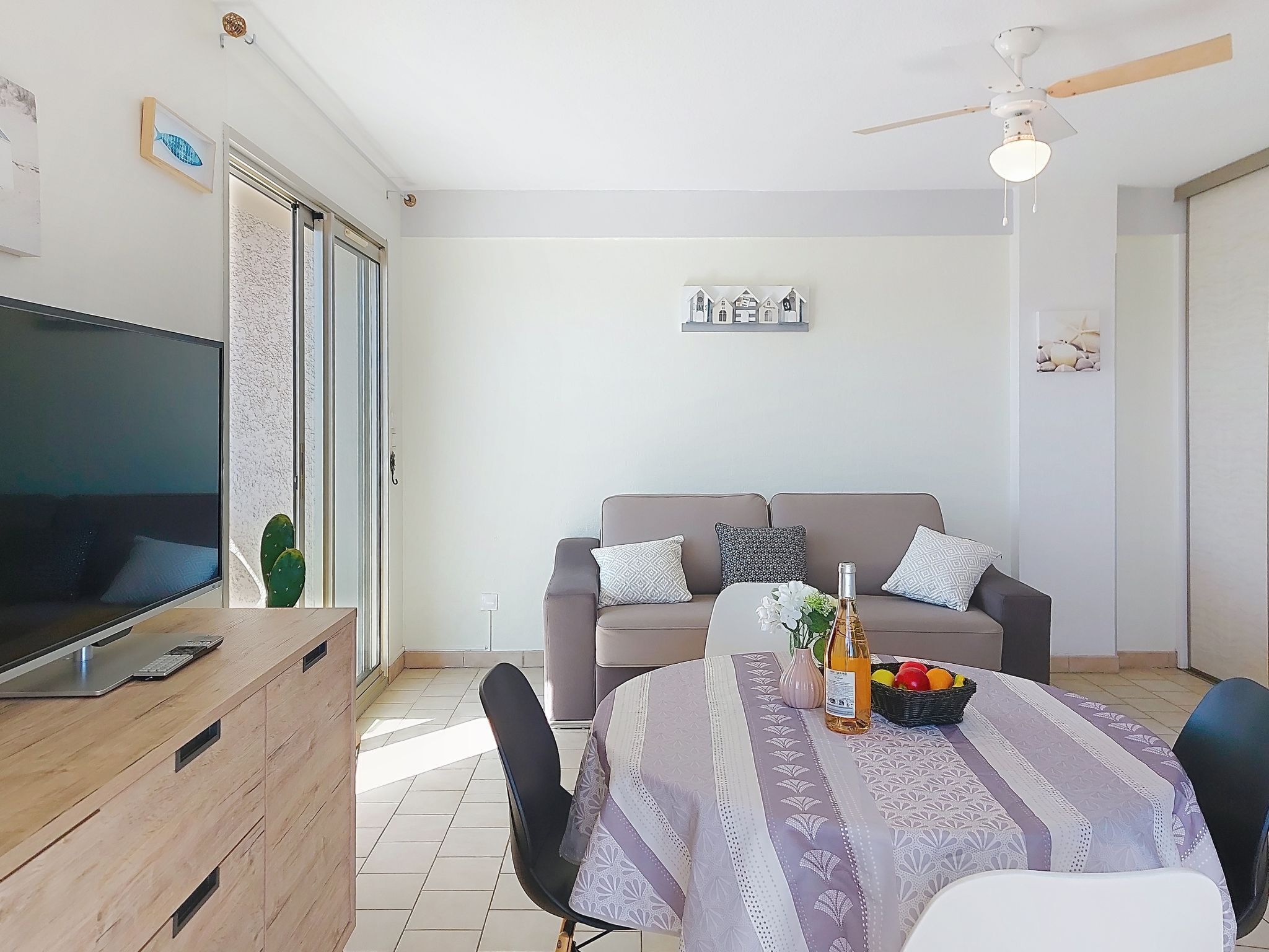 Uma sala de estar com televisão, uma mesa de jantar com toalha de mesa estampada, um sofá com almofadas padronizadas, um ventilador de teto e uma planta cacto.