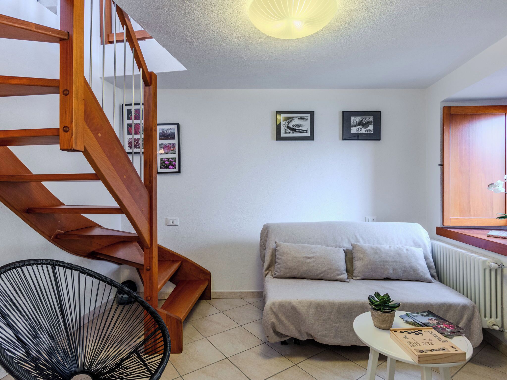 Een moderne woonkamer met een houten trap, een bank met kussens, een klein tafeltje met boeken en een plant, en ingelijste foto's aan de muur.
