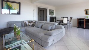 Un salon moderne avec un canapé d’angle gris, une table basse en verre et un coin repas comprenant une table et des chaises.