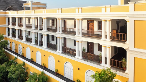 A multi-story building with a yellow facade, white columns, and balconies with wooden railings. - The Colonial Courtyard (Vanur)