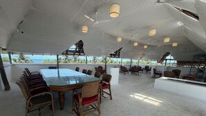 A covered outdoor dining area with a glass-top table and wicker chairs, surrounded by ceiling fans and pendant lights. - The Colonial Courtyard (Vanur)