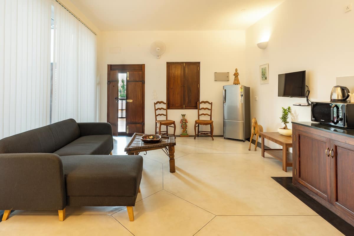 A living room with a sofa, a dining table with chairs, a kitchen area with a refrigerator, and a TV mounted on the wall.