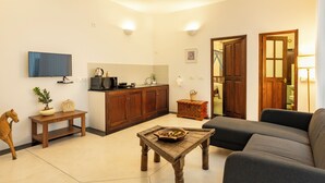 A living room with a sofa, a wooden coffee table, a television, and a kitchen area with cabinets and a microwave. - The Colonial Courtyard (Vanur)