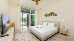 A modern bedroom with a large bed, a ceiling fan, a TV, and a view of the outdoors through a sliding glass door. - Villa Aura 3BR Private Pool Near Naiharn (Rawai)