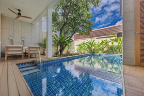 A swimming pool with a wooden deck, a ceiling fan, and a large tree in the background.