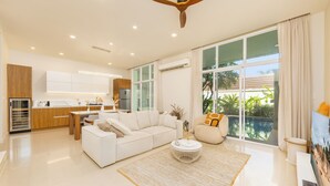 A modern living room with a white sofa, a coffee table, and a large window with a view of greenery. - Villa Aura 3BR Private Pool Near Naiharn (Rawai)