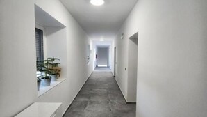 A narrow, well-lit hallway with a tiled floor and white walls. There are potted plants on a ledge on the left side. - Rooms Burnum - Twin Room 7 (Vrbnik-Knin)