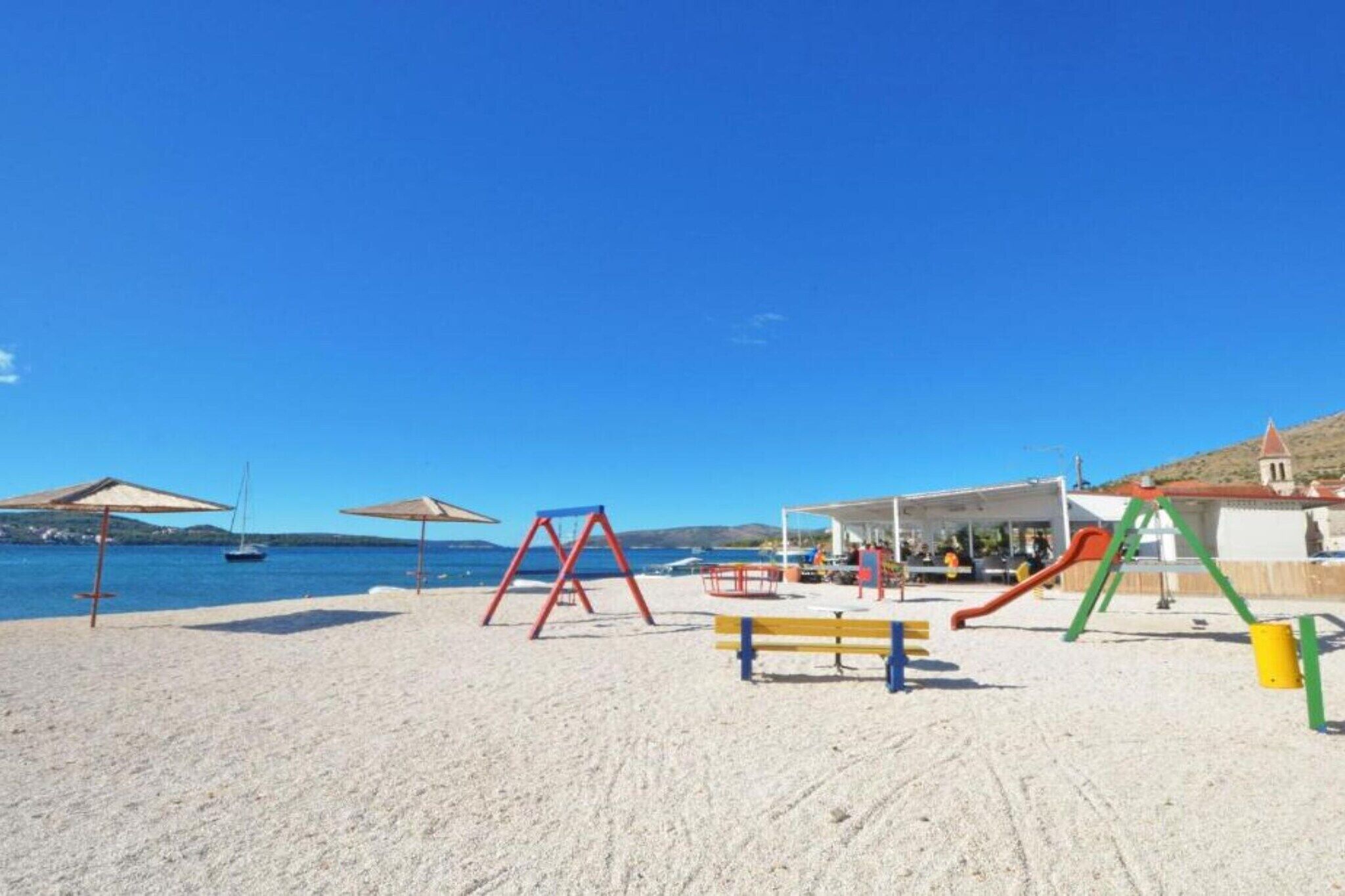 Ein Strand mit einem Spielplatz, inklusive Schaukeln, einer Rutsche und Sonnenschirmen.