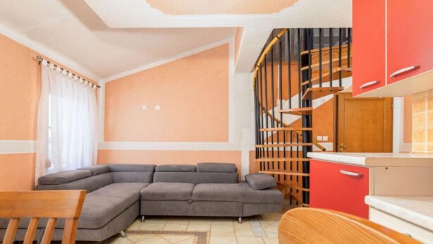 Un salon avec un canapé d’angle gris, une table à manger en bois avec des chaises, un coin cuisine avec des meubles rouges et un escalier avec une rampe en bois.
