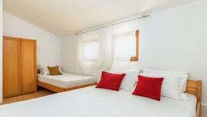 Une chambre avec un grand lit, un plus petit lit, une armoire en bois et une fenêtre ornée de rideaux légers.
