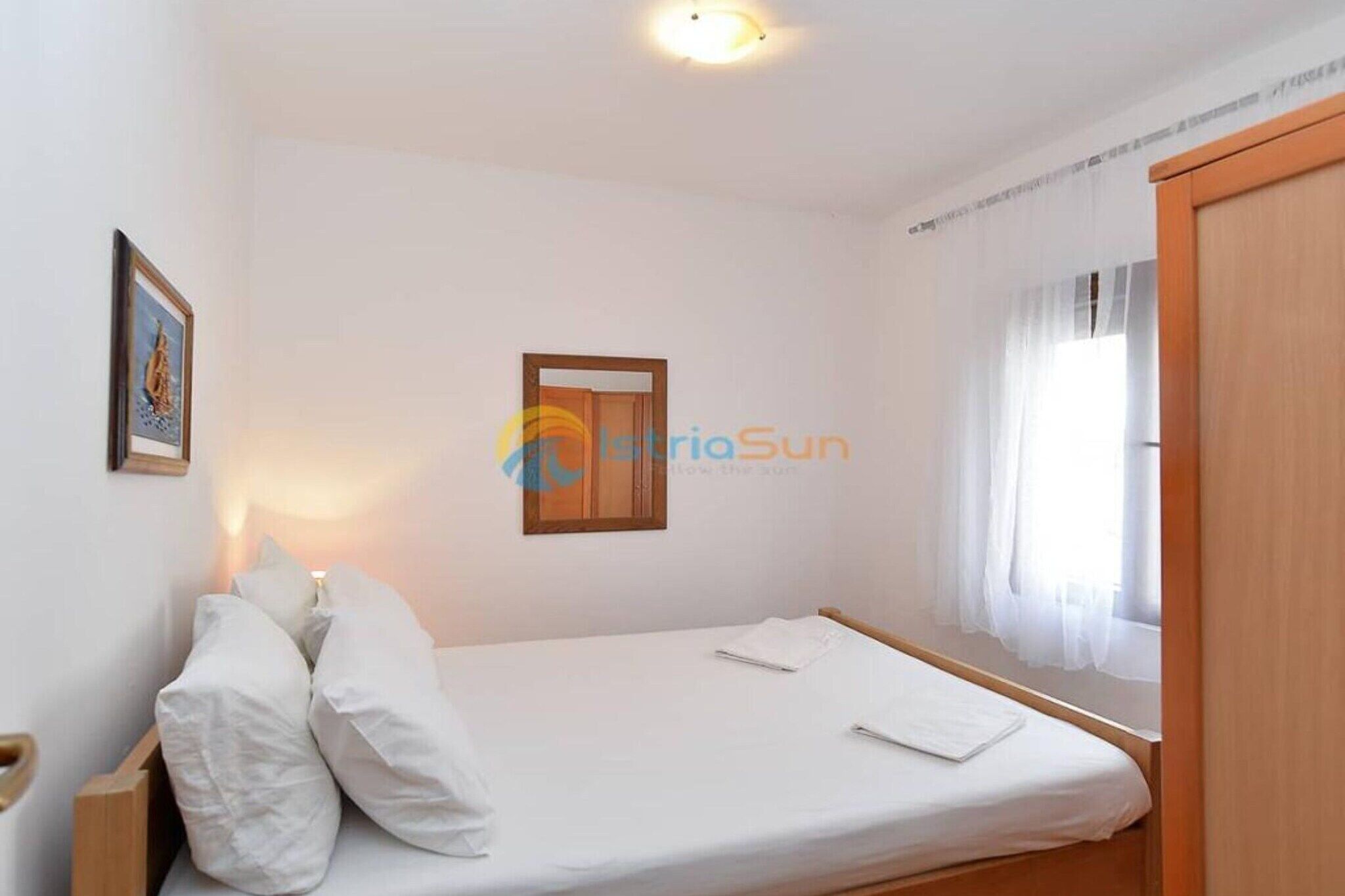 A simple, clean room with a single bed, a small window with sheer curtains, and a wooden door.