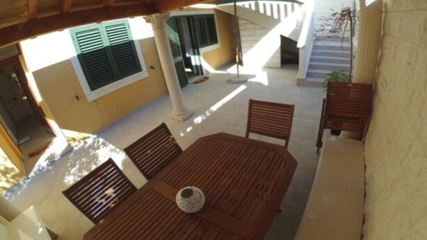A covered outdoor area with a wooden table and chairs, a white wall, and a staircase leading to an upper level.