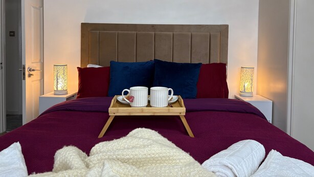 Un lit bien fait, sur lequel repose un plateau en bois portant deux tasses blanches et une théière, le tout agrémenté de coussins rouges et bleus, et flanqué de deux lampes de chevet.