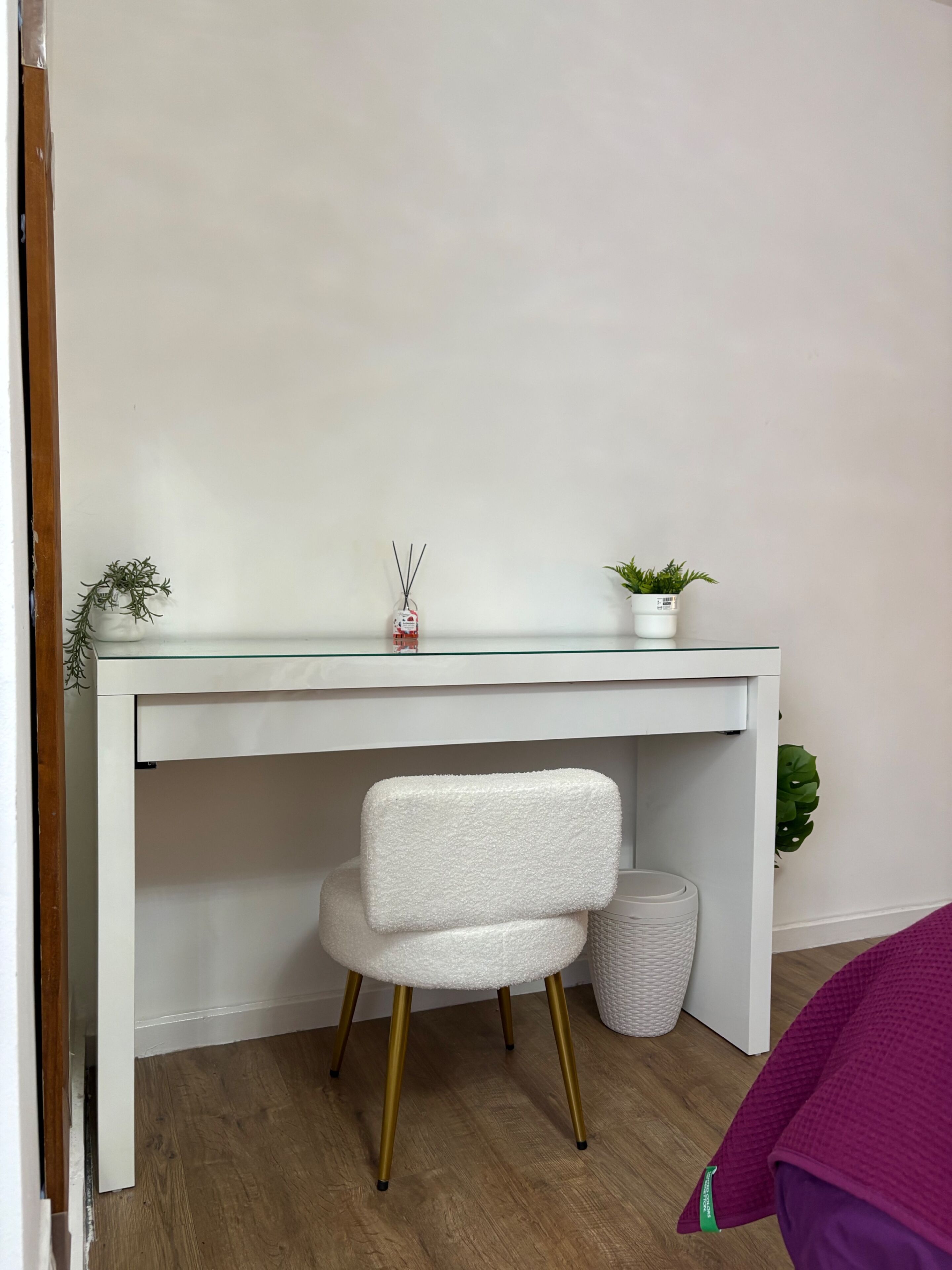 A white desk with a glass top, a white chair with gold legs, a small potted plant, and a white trash bin.