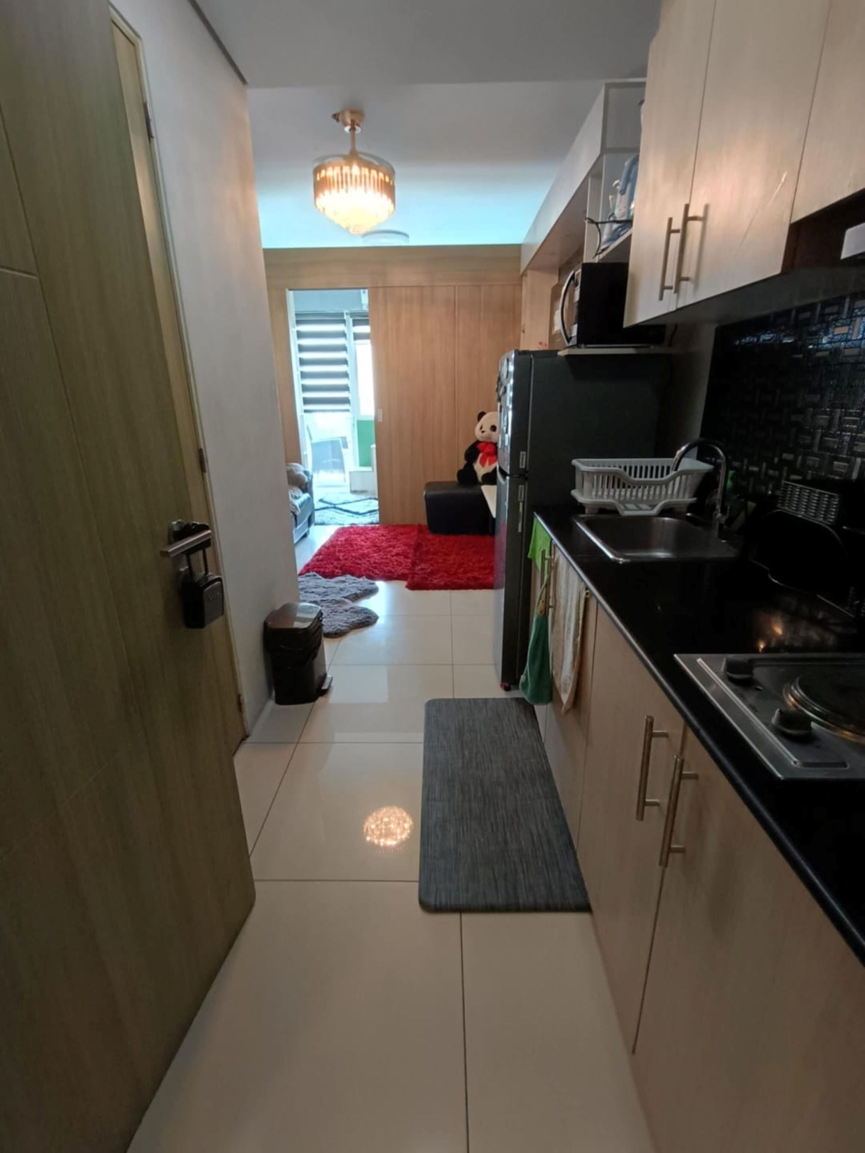 A narrow kitchen with a refrigerator, sink, and wooden cabinets. A red rug is on the floor, and a chandelier hangs from the ceiling.