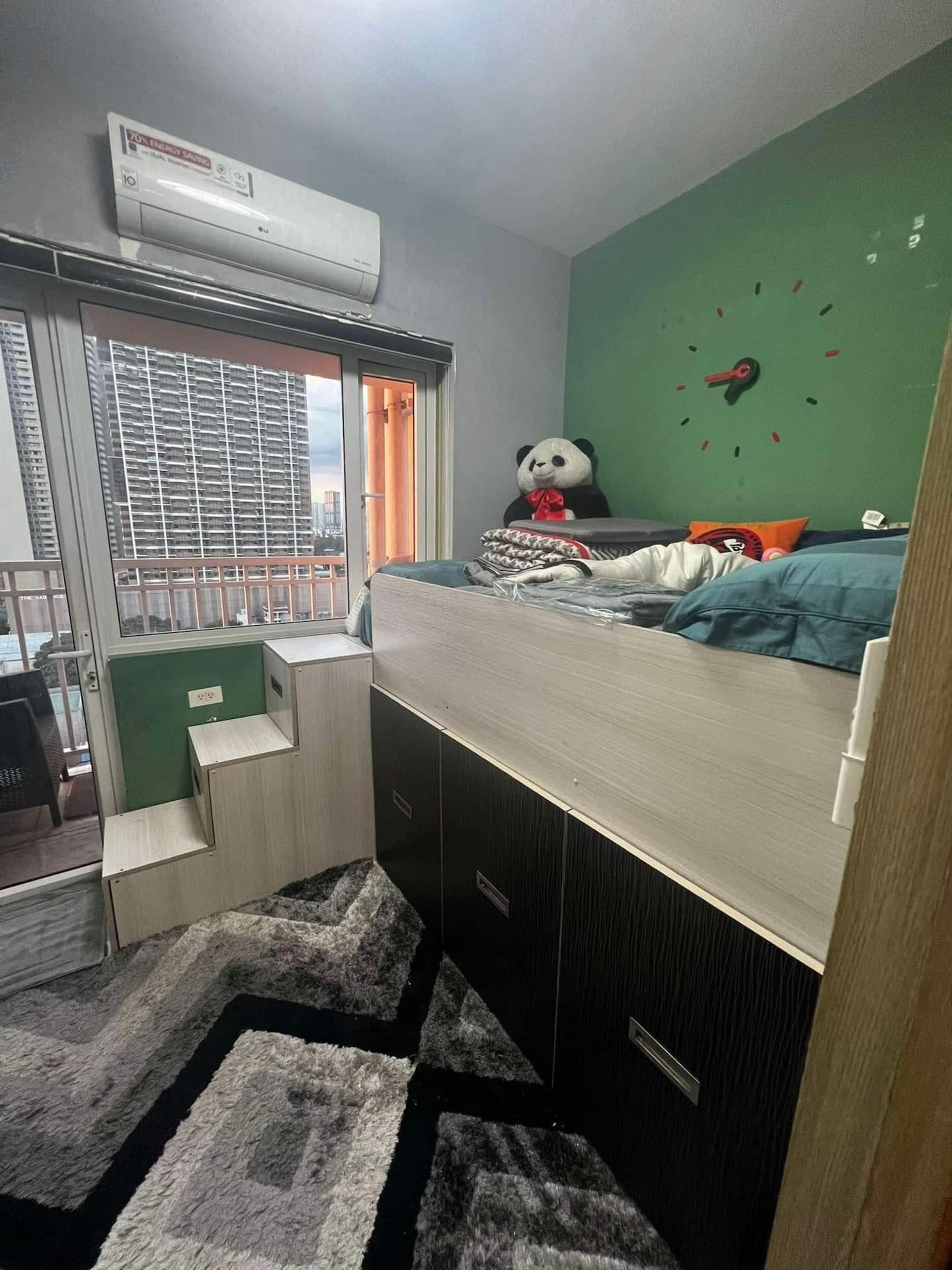 A compact room with a bed, a large panda plush toy, a green wall clock, and a window with blinds.