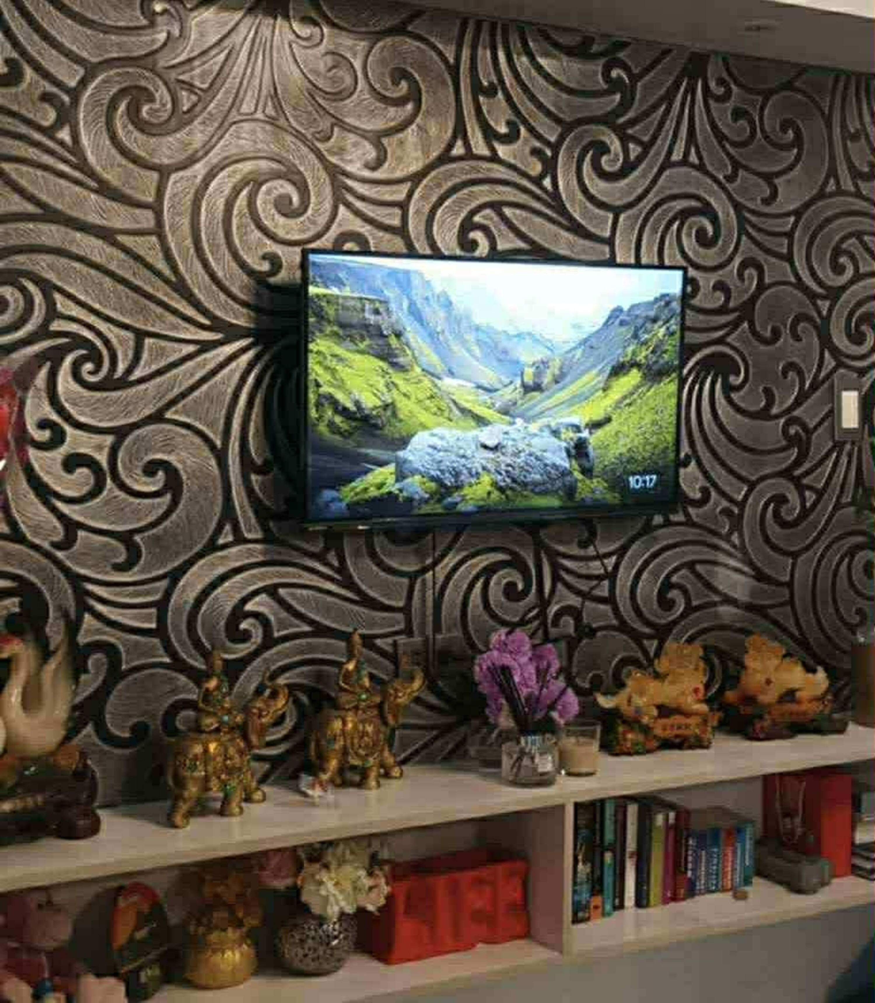Un televisor montado en la pared con un diseño decorativo, un estante con varios objetos, incluyendo figuritas y libros, y un reloj que marca las 10:17.