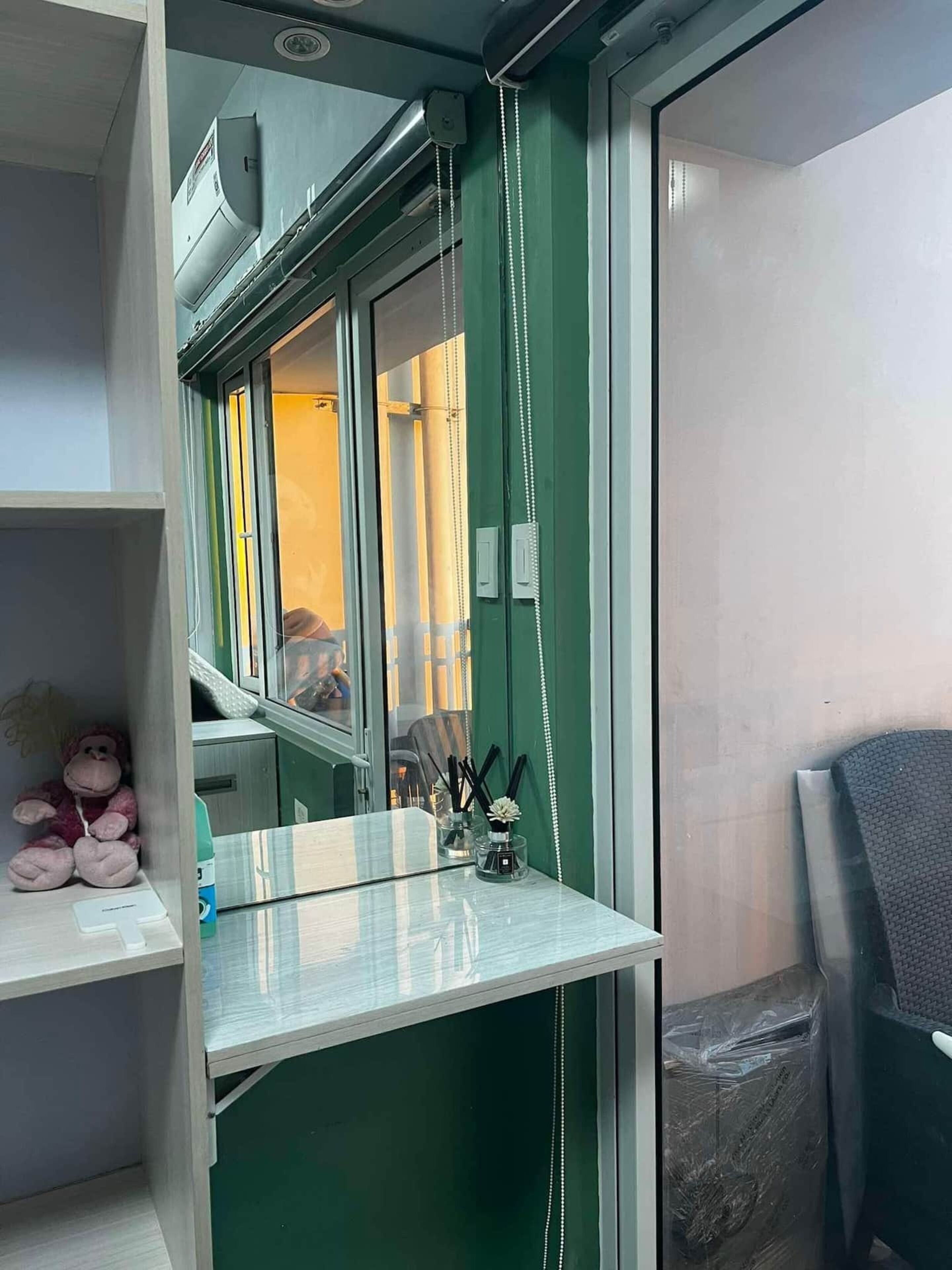 Ein kleiner, kompakter Raum mit einer grünen Akzentwand, einem Fenster mit Jalousien, einem kleinen Tisch mit Glasplatte und einem Stuhl.