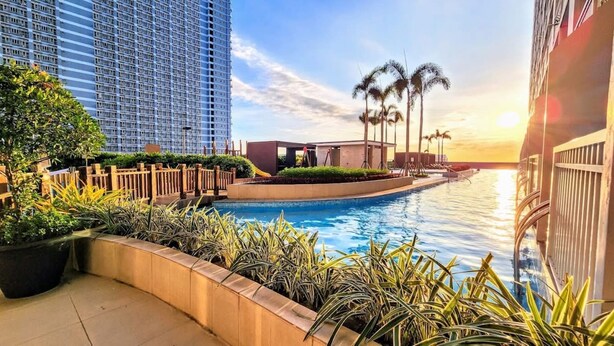 A pool area with a view of a tall building, palm trees, and a sunset. - Breeze Residences Condos (Pasay)
