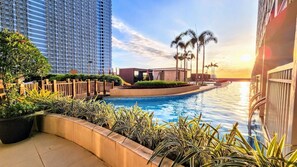 A pool area with a view of a tall building, palm trees, and a sunset. - Breeze Residences Condos (Pasay)