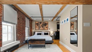 A bedroom with a bed, bench, large windows, brick walls, wooden beams, and artwork. - Two Story Penthouse - Walk to the Convention Ctr (Indianapolis)