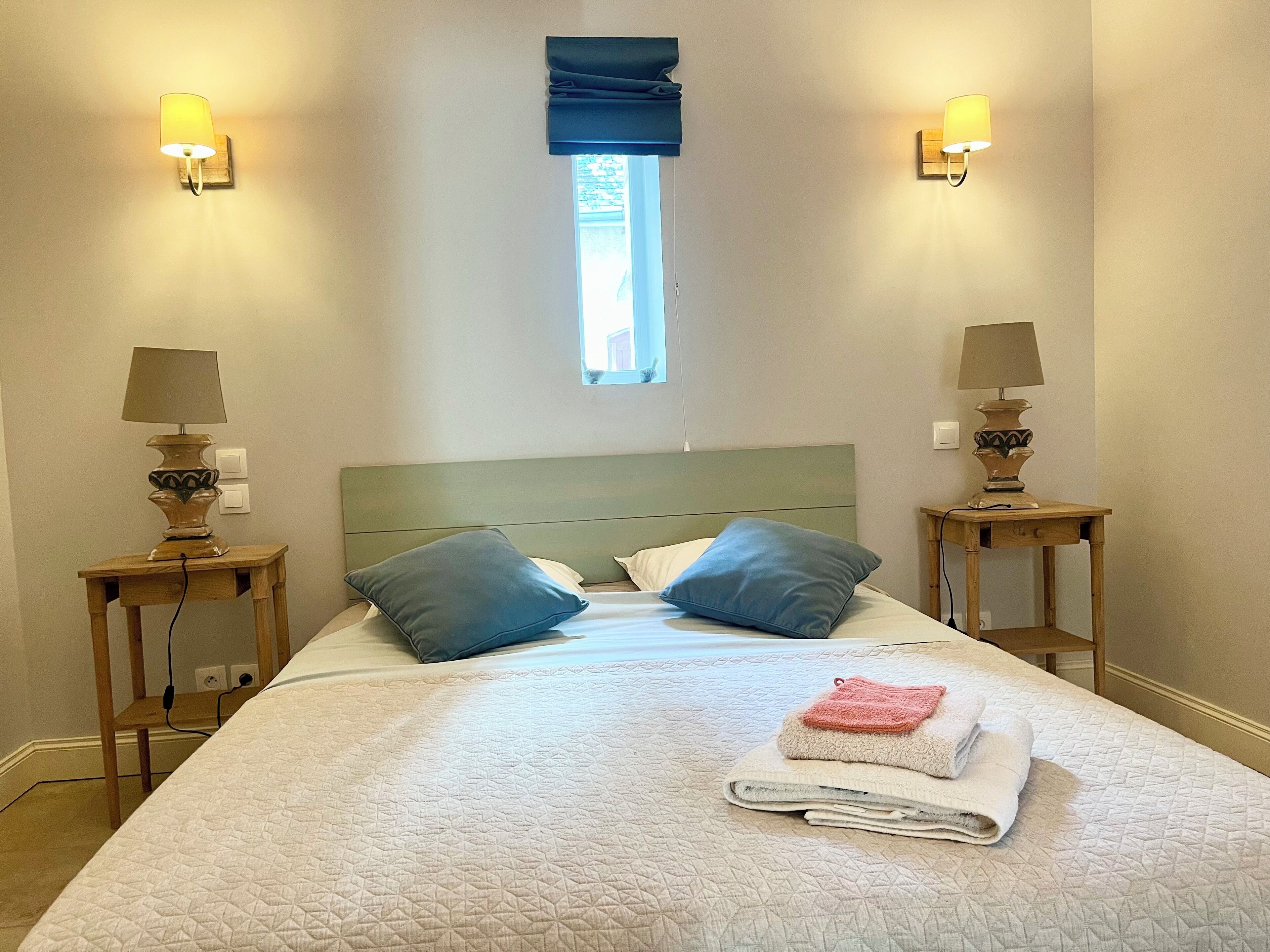 Un lit bien fait, avec une couverture matelassée, deux oreillers et une serviette pliée posée sur une table de chevet en bois.