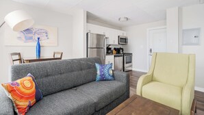 Een moderne woonkamer met een grijze bank, kleurrijke kussens, een houten salontafel en een gele fauteuil. In de achtergrond staat een eettafel met stoelen en een blauwe vaas.