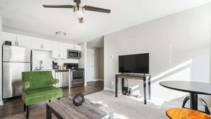 Een moderne keuken met witte kasten, een roestvrijstalen koelkast en een magnetron. Er staat een groene bekleden stoel en een houten salontafel. Een flatscreen televisie staat op een meubelstuk naast een klein rond tafeltje.