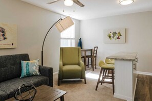 A living room with a sofa, a green armchair, a coffee table, a dining table with chairs, a floor lamp, and artwork on the walls. - Book 7 Apts Together - Near Lucas Oil Stadium (Indianapolis)
