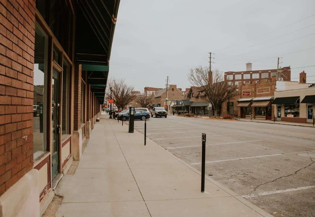 A street view with a sidewalk, parked cars, and buildings with storefronts.