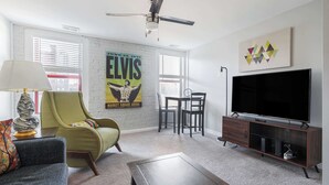 Un salon avec un fauteuil vert, un canapé, une table basse, un téléviseur posé sur un meuble en bois, un ventilateur de plafond et une affiche accrochée au mur.
