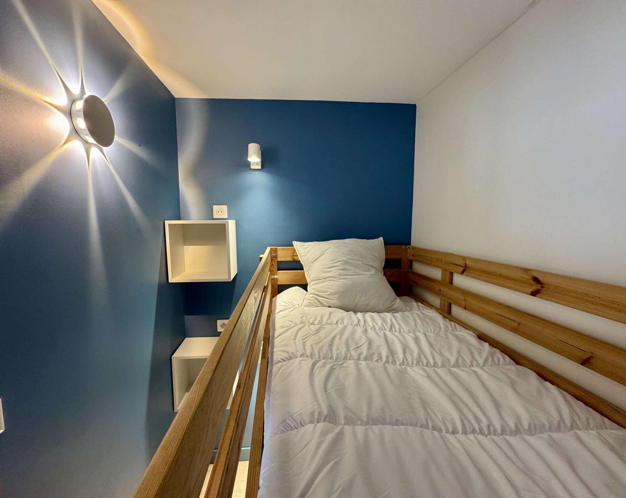 A single bed with a wooden headboard, a bedside table, a wall-mounted light, and a power outlet.
