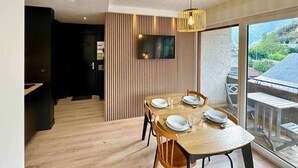 A dining area with a wooden table set for four, a TV mounted on a slatted wall, and a balcony with a view of the landscape. - New apartment for 6 people - close to the city center (Bagnères de Luchon)
