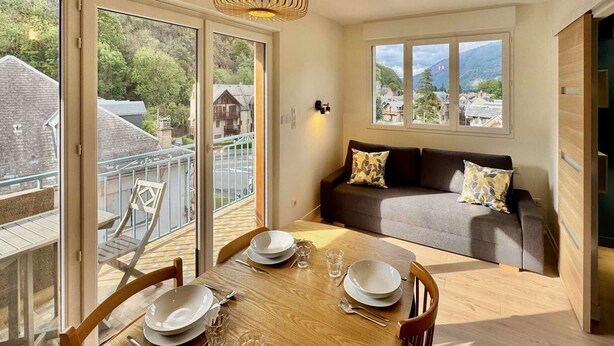 A dining area with a wooden table set for four, a sofa with cushions, and a balcony with a railing. - New apartment for 6 people - close to the city center (Bagnères de Luchon)