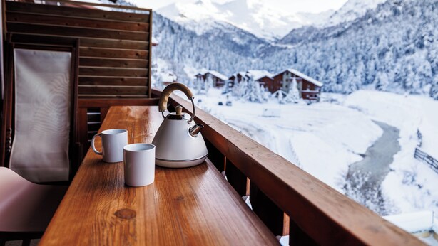 A wooden table with two white mugs and a kettle, overlooking a snowy mountain landscape with houses.