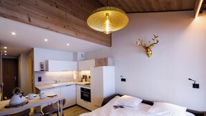 A modern hotel room with a wooden ceiling, a bed with white linens, a small kitchen area, and a decorative deer head on the wall.