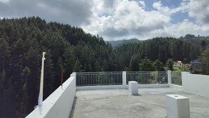 Une terrasse sur le toit, dotée d’un sol en béton, de rampes blanches et offrant une vue sur une chaîne de montagnes densément boisée, sous un ciel partiellement nuageux.
