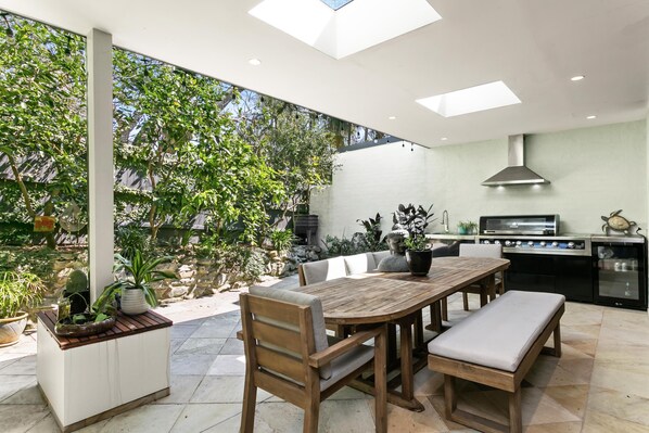 A modern outdoor kitchen with a dining area, surrounded by lush greenery and a stone wall. - Fairlight - FAIR1 - Cohen Street - Private & secluded semi in a leafy garden setting (Fairlight)