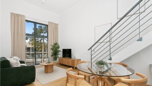 A modern living room with a glass dining table, wooden chairs, a green sofa, a TV, and a staircase.
