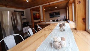 Una mesa de comedor de madera con mantel blanco, rodeada de sillas blancas. Se ve un refrigerador de acero inoxidable a la izquierda.