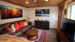 Una sala de estar con un sofá de cuero marrón, cojines rojos y beige, una mesa de centro redonda, un mueble de madera oscura y un cuadro con una escena de bosque.
