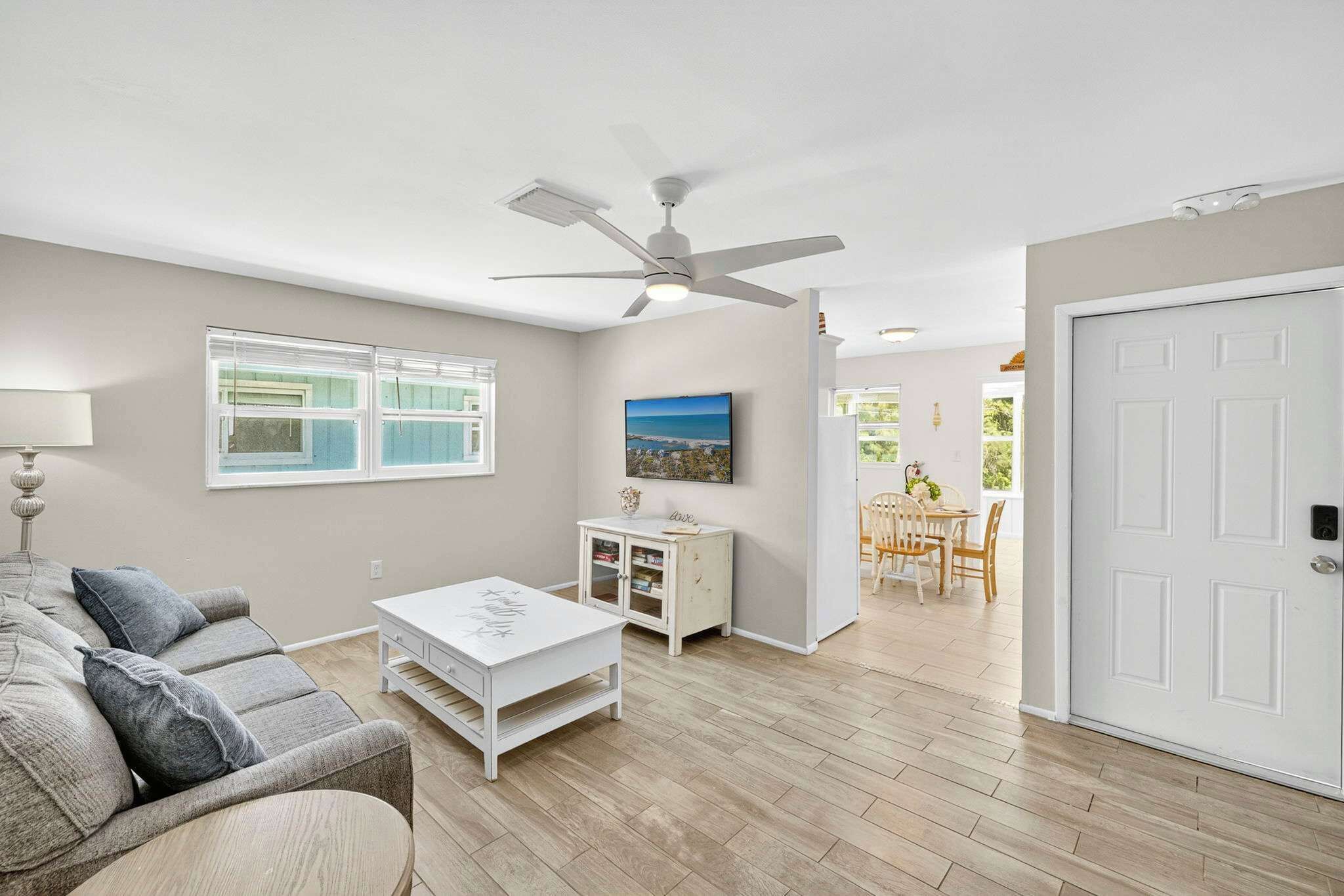 A living room with a sofa, coffee table, and a ceiling fan. There is a window with blinds, a small cabinet, and a door leading to another room.
