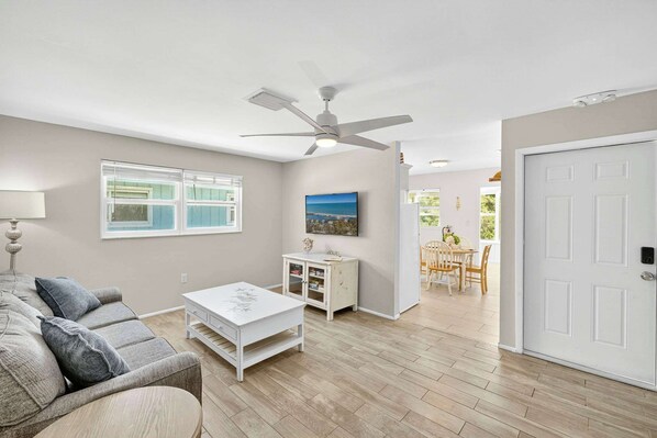 A living room with a sofa, coffee table, and a ceiling fan. There is a window with blinds, a small cabinet, and a door leading to another room.