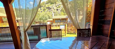 Una terraza de madera con una mesa y sillas, un espacio cubierto con cortinas translúcidas y vistas a un paisaje montañoso.