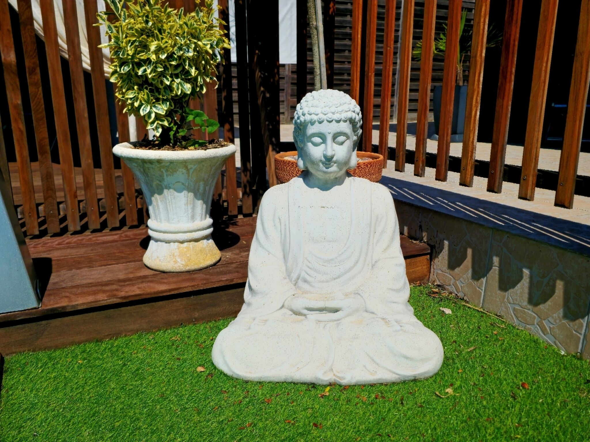 Una estatua de Buda sentada sobre césped en un jardín con una valla de madera y una planta en maceta al fondo.