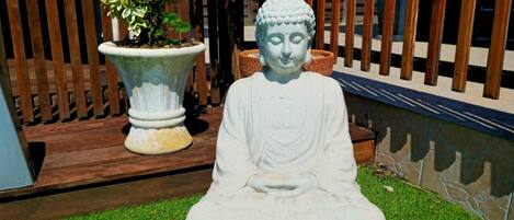 Una estatua de Buda sentada sobre césped en un jardín con una valla de madera y una planta en maceta al fondo.