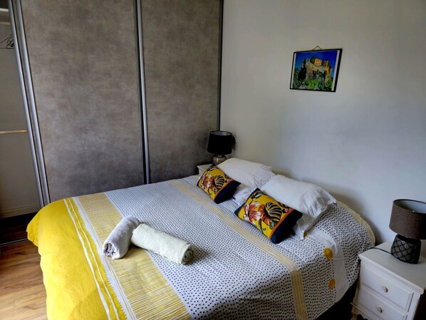Un lit bien fait, recouvert d’une couverture à rayures jaunes et blanches, avec deux oreillers à motifs floraux et une serviette blanche enroulée. Une petite table de chevet est placée à côté du lit, avec une lampe et un tableau encadré accroché au mur.