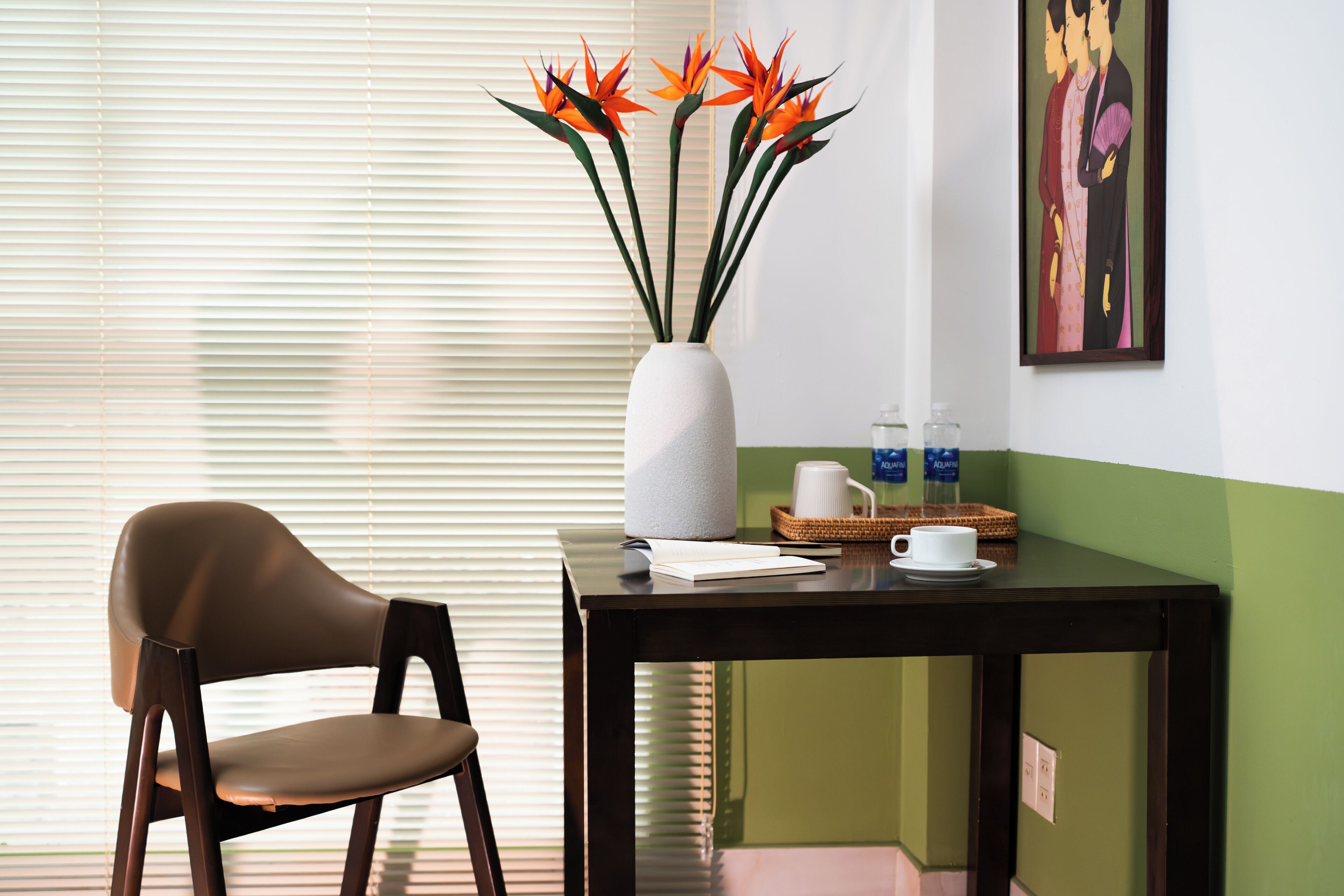 Une pièce comprenant une chaise, une table avec un vase de fleurs, un tableau et une fenêtre avec des stores.